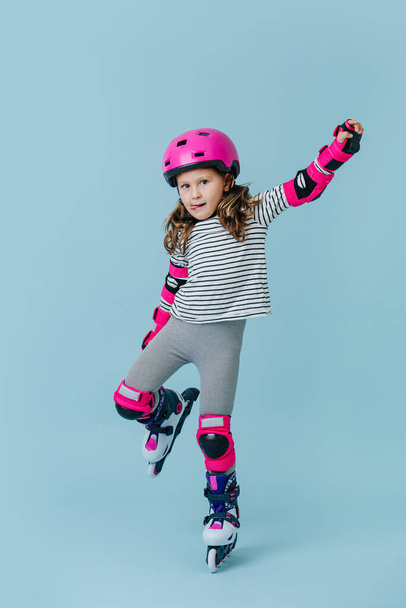 Serious focused little girl in striped clothes and roller skates with pink protective gear over blue background. She's showing off for a photo, standing on one leg, holding other one with her hand. - Photo, Image