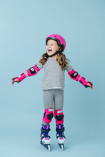 Thrilled happy little girl in striped clothes and roller skates with pink protective gear over blue background. Her arms wide apart for balance. - Photo, image