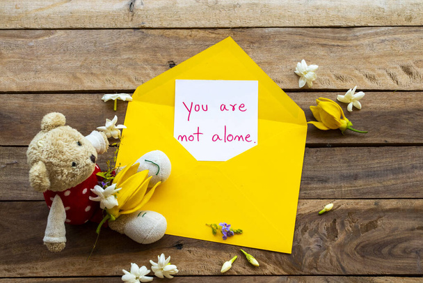 Вы не одиноки в написании посланий почерком в желтой эмали с белыми цветами жасмина, желтыми цветами ягненка и плюшевым медведем в аранжировке открытки на фоне дерева
 - Фото, изображение