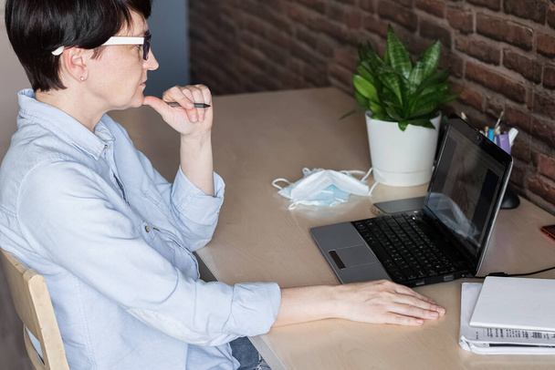 Koncepcja odległości pracy podczas wirusa. Kobieta pracuje w cyfrowym laptopie w domu, a obok którego maski medyczne. - Zdjęcie, obraz