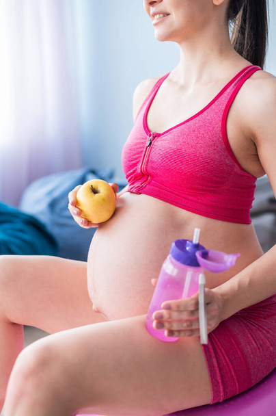 顔のない妊婦はヨガの後にリンゴを食べる。期待される母親は、ピラティスの後に果物や飲料水をかむ。フィットネスボールに座っている女の子. - 写真・画像