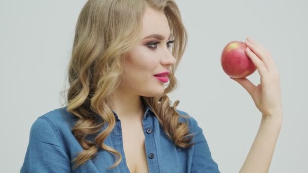 Cute woman at a table holding an apple on a background of fruit and vegetables. - Felvétel, videó