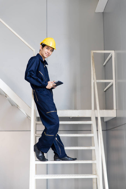Giovane e affascinante ingegnere asiatico che indossa tute blu scuro e cappello rigido di sicurezza giallo in piedi su scale metalliche in cantiere. Idee per la prevenzione e la sicurezza sul luogo di lavoro
. - Foto, immagini