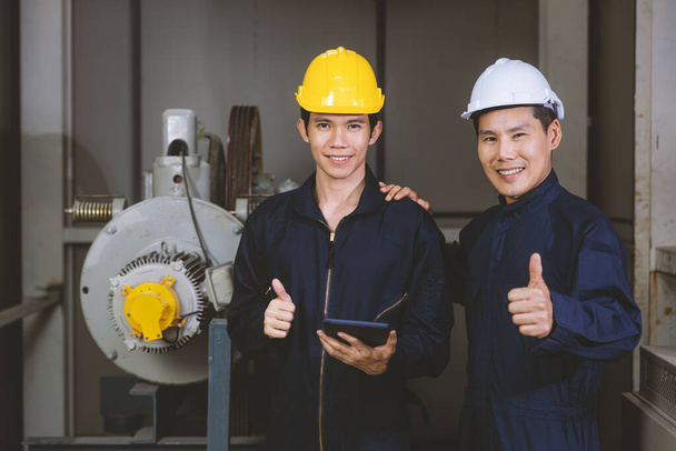 Young and handsome Asian engineers wearing dark blue coveralls and safety hard hat working together in machine room. The junior holding tablet computer and rise hands thumbs up. - Photo, Image
