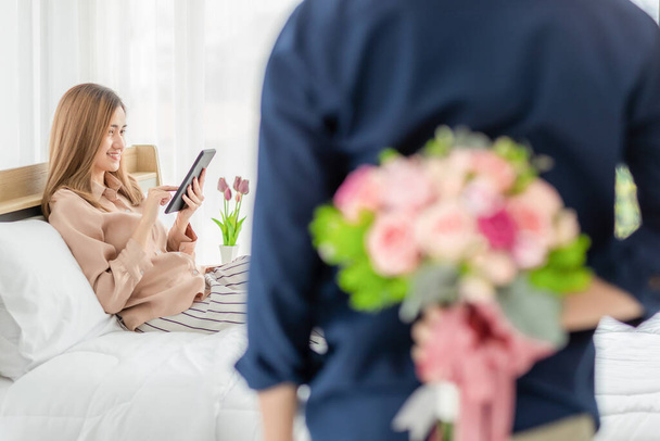 Mężczyzna ukrywający buch kolorowych kwiatów z tyłu przygotować się do dać dla kobiety, która siedzi na łóżku i gra w smartfona. Wybiórcza koncentracja na dziewczynie. Pojęcie miłości i rocznicy. - Zdjęcie, obraz