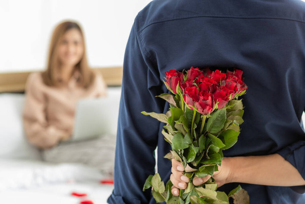 Muž skrývá spoustu barevných květin na zadní straně připravit dát pro ženu, která sedí na posteli a hraje smartphone. Selektivní soustředění na dívku. Koncept lásky a výročí. - Fotografie, Obrázek