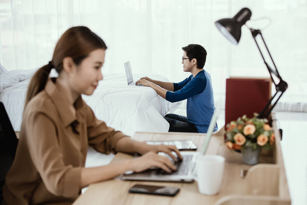 彼の妻が寝室のベッドでノートパソコンと仕事をしている間、アジアのビジネスマンは寝室のベッドの上の家から働くノートパソコンを使用しています。従業員とスタートアップのためのモダンなオフィスホームベースの仕事のためのアイデア. - 写真・画像