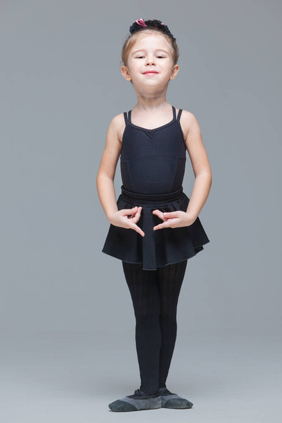 Beautiful little girl gymnast in black sportswear is going to become ballet dancer and shows exercises on grey background - Photo, Image