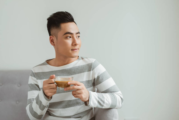 自宅でソファに座っているときにアジアの男はコーヒーを飲む. - 写真・画像