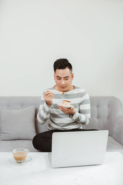 Jonge Aziatische man die taart eet als hij op de bank zit en laptop gebruikt. Werk vanuit huis. - Foto, afbeelding