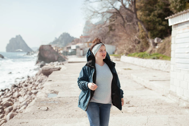 A pregnant happy woman walks along the promenade along the spring sea, she is dressed in a jacket and jeans and smiling - Foto, imagen