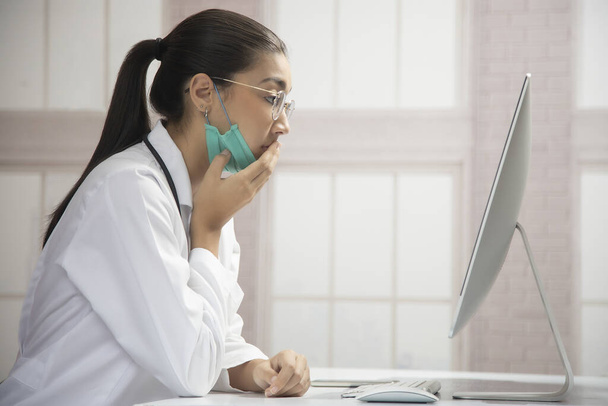 молодая женщина в маске в платье врача с рукой на губах, глядя на компьютер
 - Фото, изображение