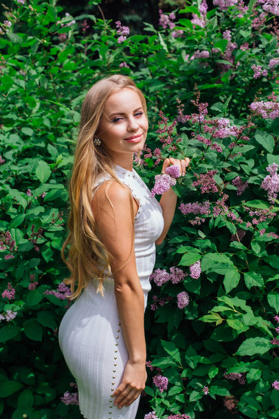 Spring portrait of a charming blond woman wearing beautiful white dress standing next to blooming purple lilac bush. - Foto, imagen