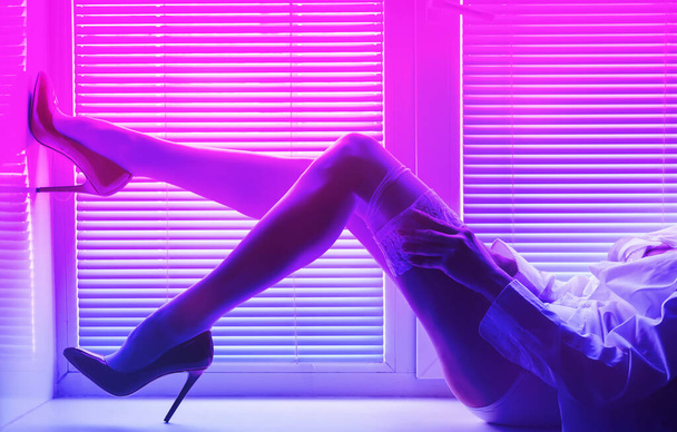 beautiful woman with long legs in a white shirt, stockings high-heeled shoes on the windowsill by the window with shutters in neon lighting - Photo, image