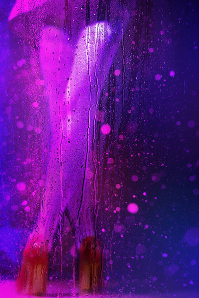 beautiful blurry female legs in neon light behind wet glass with raindrops - Valokuva, kuva
