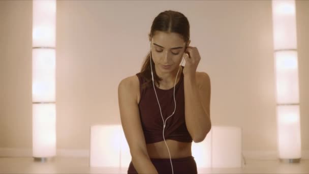 Girl sitting in lotus pose on mat. Woman in earphones using smartphone in studio - Footage, Video