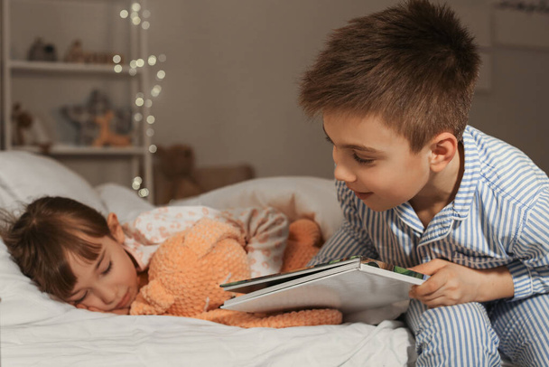 Little girl falling asleep while her brother reading bedtime story at home - 写真・画像