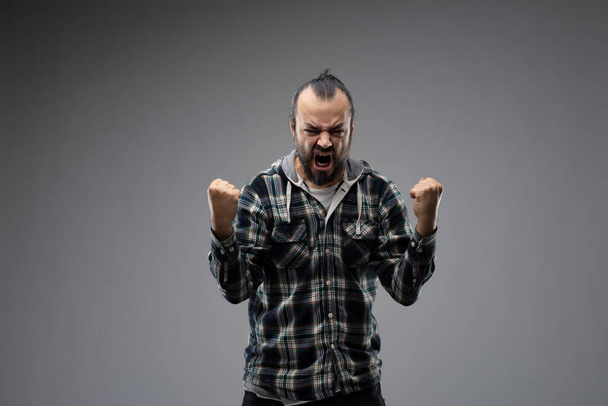 Een man met een baard in een geruit shirt die agressief zijn succes toont met een winnend gebaar, staande tegen een grijze achtergrond met gebalde vuisten en schreeuwend Ja. Voorportret met kopieerruimte - Foto, afbeelding