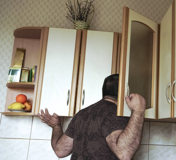 Young man in shirt peeking  into kitchen cupboard at home, viewed from the back side. alone hungry male bachelor search any food.  - Фото, зображення