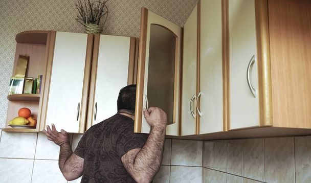 Young man in shirt peeking  into kitchen cupboard at home, viewed from the back side. alone hungry male bachelor search any food.  - Фото, зображення