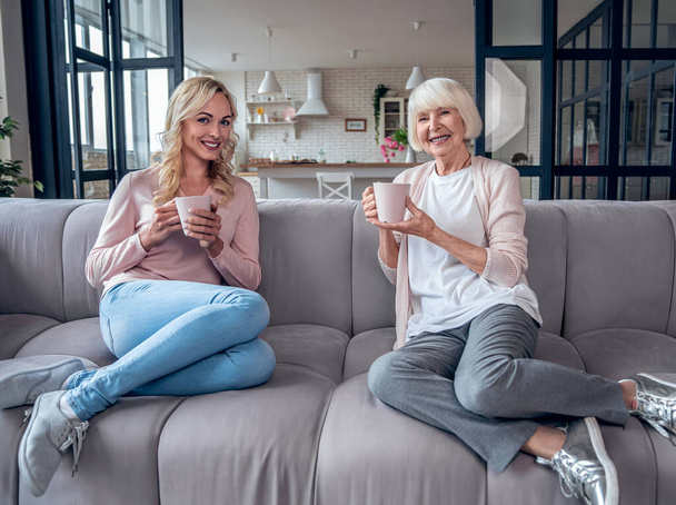 Старшая женщина и ее привлекательная дочь проводят время вместе дома. Сидеть на диване и пить чай вместе. Happy Mothers 'Day
 - Фото, изображение