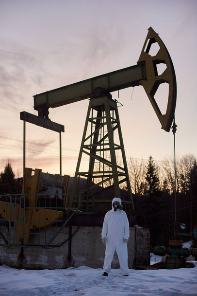 Pełna długość męskiego naukowca w masce gazowej stojącego na terytorium pola naftowego z pompą olejową rocker-machine na tle - Zdjęcie, obraz