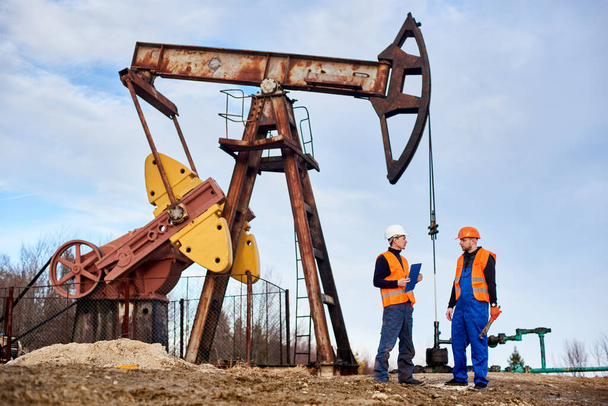 Δύο μηχανικοί πετρελαίου σε κράνη και γιλέκα εργασίας στέκεται στο έδαφος του κοιτάσματος πετρελαίου με την αντλία πετρελαίου και τον ουρανό στο παρασκήνιο. Πετρέλαιο άνθρωπος κρατώντας πρόχειρο και μιλώντας με συνάδελφο στον τομέα του πετρελαίου. - Φωτογραφία, εικόνα