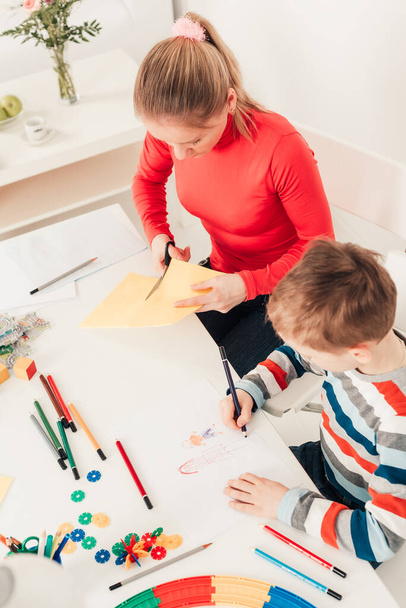 Mãe com o filho recortando papel e desenhando em conjunto na mesa branca
 - Foto, Imagem