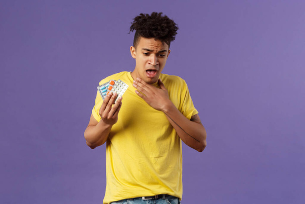 Health, influenza, covid-19 concept. Portrait of young man coughing, choking with tablet, drank pill without water, holding drugs and touch neck as suffer sore throat, purple background - Photo, Image