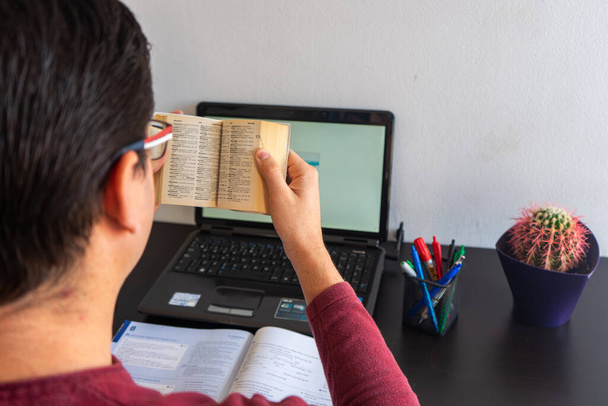 Człowiek uczący się angielskiego. Ma w ręku słownik. Na biurku jest podręcznik, laptop i kaktus.. - Zdjęcie, obraz