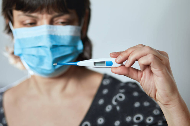 Woman in a mask with Coronavirus reads his temperature on a thermometer. Female measures the temperature. Check body temperature for virus symptoms. Copy space. Coronavirus. 2019 Novel Coronavirus (2020-nCoV) - Foto, imagen