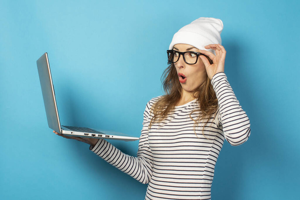 νεαρή κοπέλα σε ένα λευκό καπέλο κοιτάζει σοκαριστικά σε ένα φορητό υπολογιστή και κρατά τα γυαλιά σε ένα μπλε φόντο. - Φωτογραφία, εικόνα