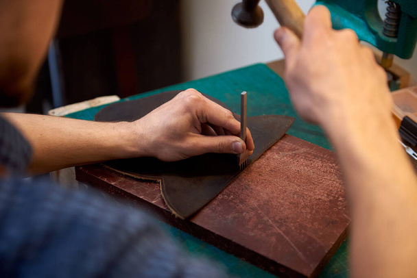 The craftsman works with a hammer and an awl to punch small holes on genuine leather for a seam. Closeup of leatherman working with brown natural leather using craft tools. - Φωτογραφία, εικόνα