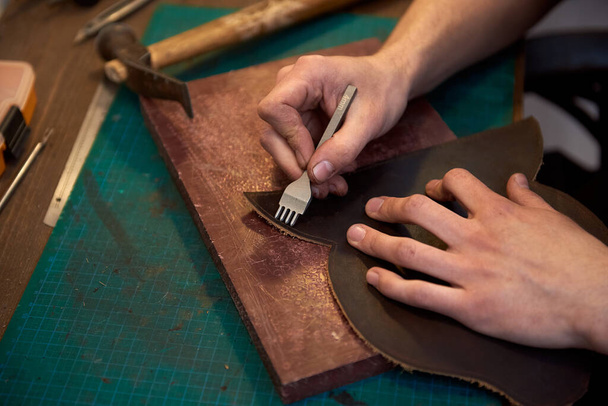 The leatherman holding crafting tool and working with brown genuine leather. Closeup of skinner craftsman working with natural leather using craft tools. Working with genuine leather. - Φωτογραφία, εικόνα