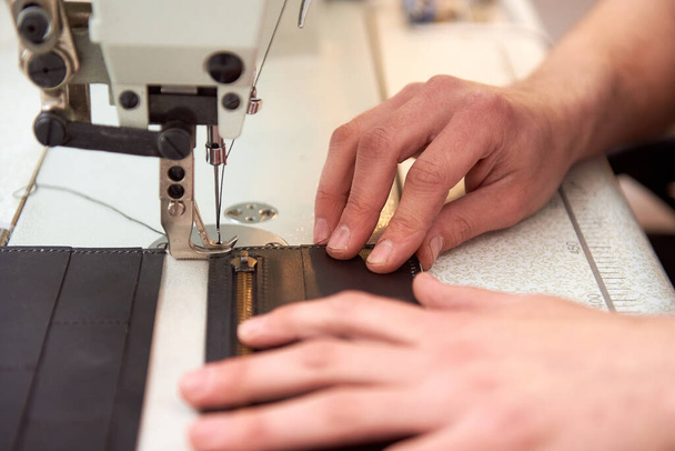 The leather craftsman produces leather products on sewing machine in his workshop. Male hands stitching gray leather wallet on white sewing machine. Manufactory and small business of leatherman. - 写真・画像