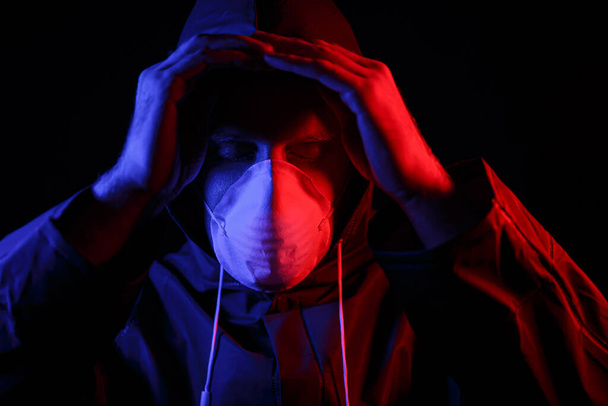 Ένας άντρας με προστατευτική λαστιχένια στολή και λευκή ιατρική μάσκα. Προστασία από ιούς. Φωτισμένο σε κόκκινο και μπλε. - Φωτογραφία, εικόνα
