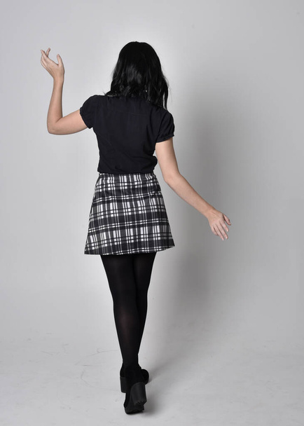 Portrait of a goth girl with dark hair wearing blue and plaid skirt with boots. Full length standing pose with back to the camera on a studio background. - Foto, immagini