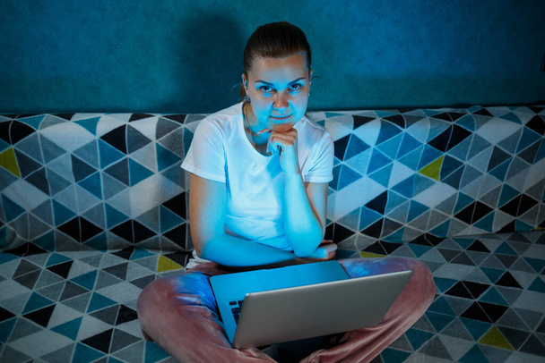 Een meisje in een wit T-shirt zit thuis op de bank met een laptop op schoot en kijkt naar het scherm. Leest nieuwe belangrijke informatie. Zoeken Nieuws - Foto, afbeelding