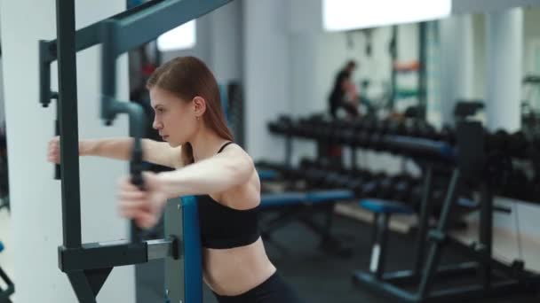 A strong young woman performs an exercise on the simulator for back and triceps. Blurred Background. Camera motion to right. Close-up - Πλάνα, βίντεο