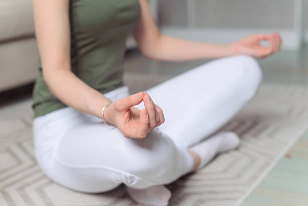 Woman does sports at home. White leggings and green top. Yoga, pilates, breathing, meditating, doing Ardha Padmasana exercise, Half Lotus pose with mudra gesture. Close up hands. Morning charging, workout, lifestyle.Well being, wellness concept. - Foto, imagen