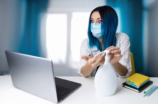 Mulher preocupada com cabelos azuis em máscara protetora olhando no conteúdo do laptop sentado no escritório em casa. pandemia do mundo do pânico viral. Fique em casa conceito
. - Foto, Imagem