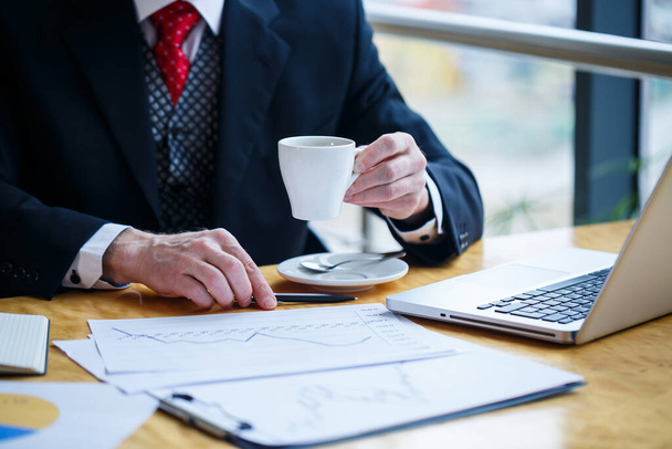 Ein erwachsener männlicher Geschäftsmann arbeitet an einem neuen Projekt und betrachtet Aktienwachstumsdiagramme. Sitzt an einem großen Fenster am Tisch. Blickt auf den Laptop-Bildschirm und trinkt Kaffee. - Foto, Bild