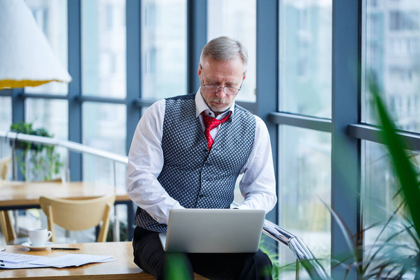 Adult male businessman, teacher, mentor working on a new project. Sits by a large window on the table. He looks at the laptop screen. - Photo, image
