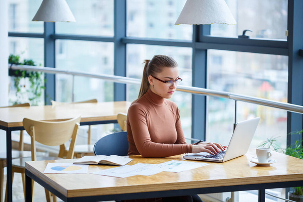 Επιτυχημένη γυναίκα διευθυντής στο δικό της γραφείο με μεγάλα παράθυρα κάνει ένα νέο επιχειρηματικό σχέδιο οικονομικής ανάπτυξης σε ένα φορητό υπολογιστή με ένα φλιτζάνι καφέ. Μια επιχειρηματίας με έγγραφα κάθεται σε ένα ξύλινο τραπέζι - Φωτογραφία, εικόνα