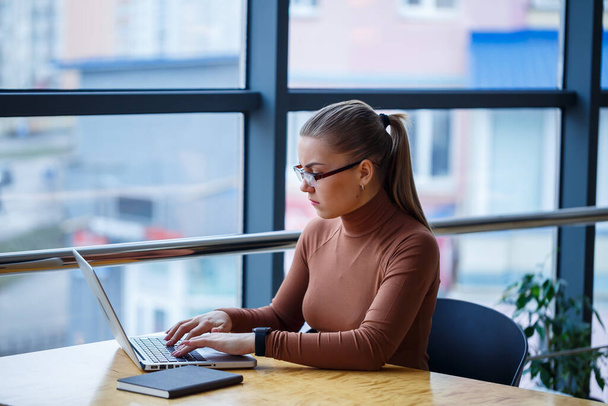 大きな窓が付いている彼女自身のオフィスの成功した女の子のマネージャーはコーヒーのコップが付いているノートパソコンの新しい経済開発のビジネス計画を作る。木製のテーブルには書類を持った女性が座っている。 - 写真・画像