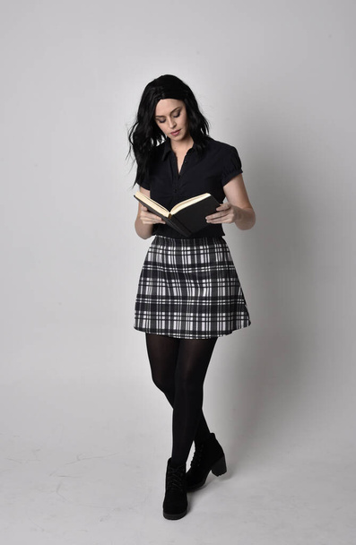 Portrait of a goth girl with dark hair wearing blue and plaid skirt with boots. Full length standing pose, holding a book, on a studio background. - Photo, Image