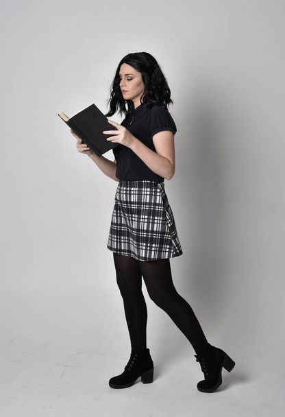 Portrait of a goth girl with dark hair wearing blue and plaid skirt with boots. Full length standing pose, holding a book, on a studio background. - Photo, Image