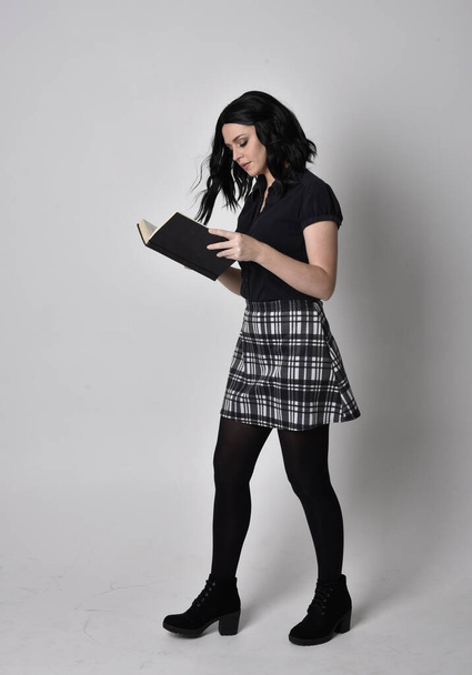 Retrato de una chica gótica con el pelo oscuro vistiendo falda azul y a cuadros con botas. Posada de pie, sosteniendo un libro, sobre un fondo de estudio
. - Foto, imagen