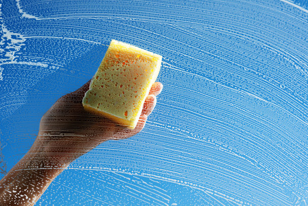 Миття вікна жовтою губкою та миючим засобом. Жіноча рука натирає мильне скло на сонячно-блакитне небо. Концепція прибирання весняного будинку
 - Фото, зображення
