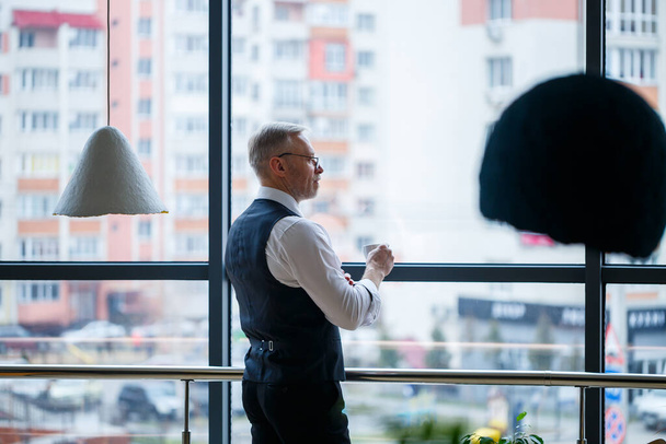 Lächelnd denkt der Geschäftsführer an seine erfolgreiche berufliche Entwicklung, während er mit einer Tasse Kaffee in der Hand in seinem Büro vor einem Fenster mit Kopierraum steht. - Foto, Bild